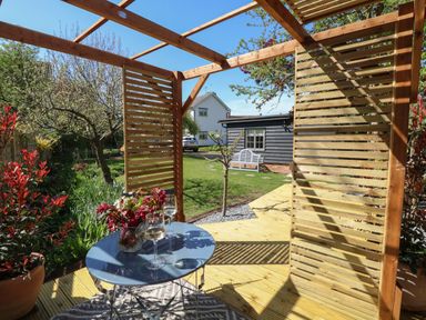 An outdoor wooden pergola with a round table holding two wine glasses and flowers with a garden and houses in the background at The Forge in Old Newton near Stowmarket