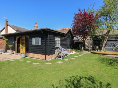 A black wooden garden shed with small windows and a door surrounded by grass and trees at The Forge in Old Newton near Stowmarket