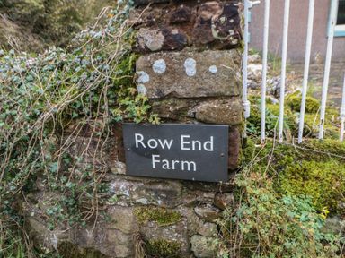 A stone wall with a Row End Farm sign surrounded by moss and plants near a white metal fence at Row End farm in Brough