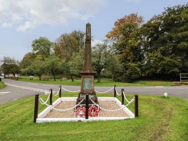 A war memorial stone monument surrounded by chains and posts with wreaths in a grassy area near a road at Row End farm in Brough