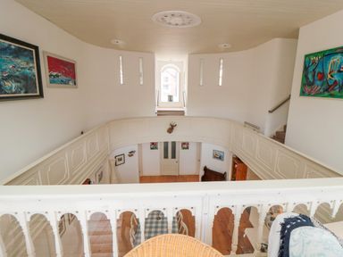 An upper level hallway with white banisters overlooking a lower room with wooden floors and framed paintings at Bethel Chapel in Staithes