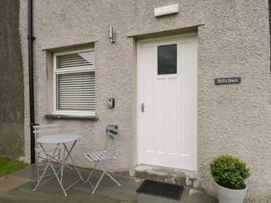 A white door and window with blinds outside with a small table and two chairs on a stone patio and a potted plant at Bill's Barn in Oxen Park near Ulverston