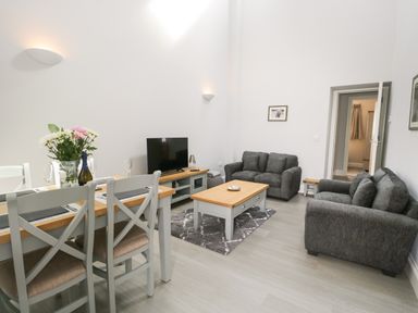 A living room with a gray sofa set a wooden coffee table a television on a wooden stand and a dining table with chairs and flowers at Bill's Barn in Oxen Park near Ulverston