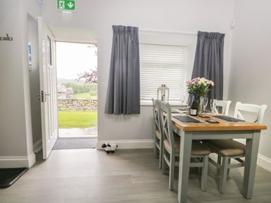 A dining area with a wooden table and chairs set with placemats glasses a flower vase and a bottle next to a window with curtains and an open door showing an outdoor stone wall and greenery at Bill's Barn in Oxen Park near Ulverston