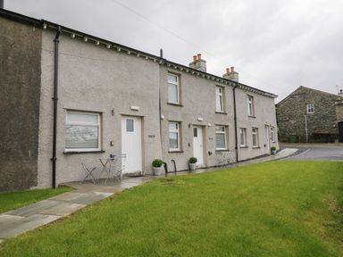 A row of connected houses with white doors and windows and small outdoor tables and chairs on a paved path with a grassy area at Bill's Barn Oxen Park near Ulverston