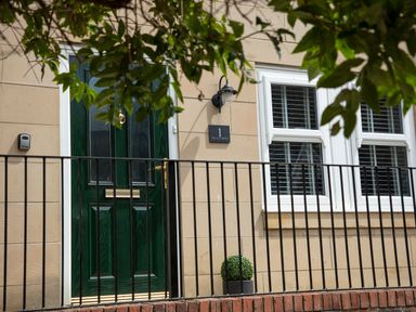 A green door next to two white windows with black railing and a small plant at 1 Wesley Mews in Alnwick