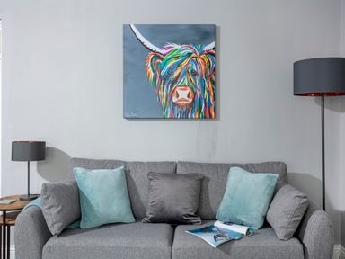 A grey sofa with cushions and an open magazine below a colorful cow painting with two lamps on either side at 1 Wesley Mews in Alnwick