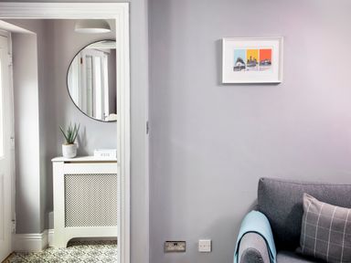 A corner of a living room with a gray couch and a blue blanket next to a hallway with a white cabinet topped with a plant and a round mirror at 1 Wesley Mews in Alnwick