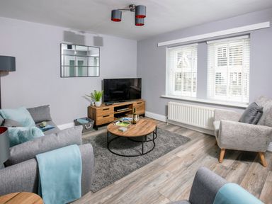A living room with gray sofas a wooden coffee table a television on a wooden stand and a window with white blinds at 1 Wesley Mews in Alnwick