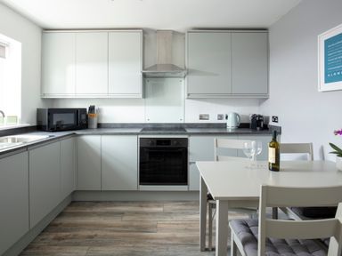 A kitchen with grey cabinets a black microwave a knife block a kettle and coffee machine and a white dining table with chairs and wine bottles and glasses at 1 Wesley Mews in Alnwick