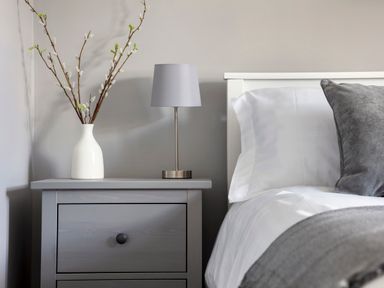 A bedside table with a lamp and a vase with branches next to a bed with pillows at 1 Wesley Mews in Alnwick