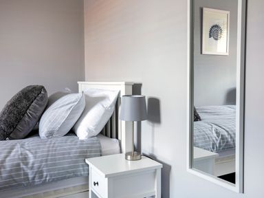 A bedroom with a bed and three pillows next to a nightstand with a lamp and a wall mirror at 1 Wesley Mews in Alnwick