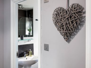 A bathroom with a sink soap dispenser mirror and a small plant seen through a doorway with a woven heart decoration on the wall at 1 Wesley Mews in Alnwick