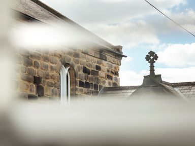 A stone building with an arched window and a roof cross under a partly cloudy sky at 1 Wesley Mews in Alnwick