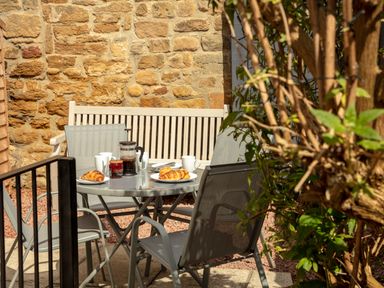 An outdoor patio with a round table set with coffee mugs croissants and a French press surrounded by chairs and a wooden bench at 1 Wesley Mews in Alnwick
