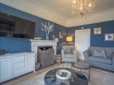 A living room with a grey sofa and armchair a white fireplace and a wall mounted TV at Argoed in Abersoch