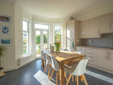 A kitchen with a wooden dining table and white chairs near windows and cabinets at Argoed in Abersoch