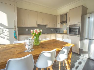 A kitchen with beige cabinets a wooden dining table white chairs a vase with flowers and glasses at Argoed Abersoch