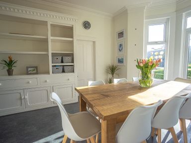A dining room with a wooden table white chairs a built in white cabinet and tulips in a vase on the table at Argoed in Abersoch