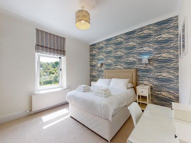 A bedroom with a bed towels on bed window desk chair and patterned wall at Argoed in Abersoch