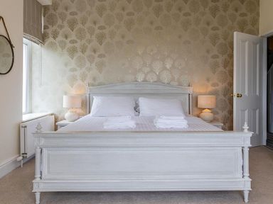 A bedroom with a large white wooden bed two bedside tables with lamps and patterned wallpaper at Argoed in Abersoch