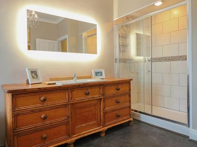A bathroom with a wooden vanity cabinet under a lit mirror and a glass door shower enclosure at Argoed in Abersoch