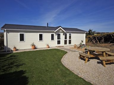An exterior view of a single-story building with a grass lawn a gravel path and a wooden picnic table in the yard at Arwelfa Lodge in Abersoch
