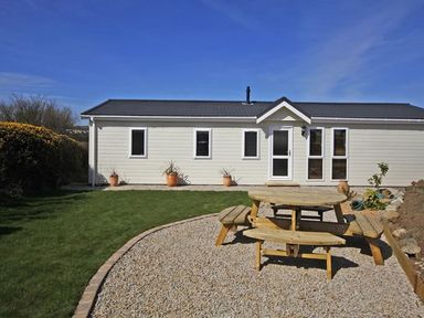 A garden with a wooden picnic table on a gravel area and a white house with multiple windows at Arwelfa Lodge in Abersoch
