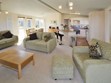 A living room with green sofas a wooden coffee table and an open kitchen with bar stools at Arwelfa Lodge in Abersoch