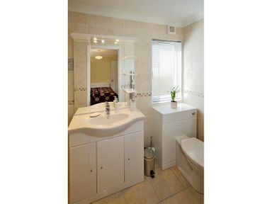 A bathroom with a sink cabinet, mirror, toilet, window with blinds, and a small plant at Arwelfa Lodge in Abersoch