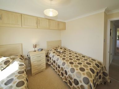 A bedroom with two single beds a nightstand with a lamp and overhead cabinets at Arwelfa Lodge in Abersoch
