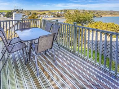 An outdoor wooden deck with a glass table and four chairs overlooking a coastal village and the sea at Arwelfa Lodge in Abersoch