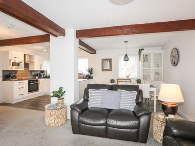 An open living space with a black leather sofa, two round wooden side tables with lamps, a dining table with white chairs, and a kitchen with white cabinets and appliances at Bywyd Da (232 Cae Du) in Abersoch