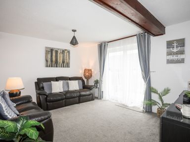 A living room with brown leather sofas, a lamp, plants, curtains, and wall art at Bywyd Da (232 Cae Du) in Abersoch