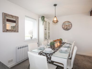 A dining room with a glass table set for eight with white chairs a wall clock a wooden framed mirror and a cabinet at Bywyd Da 232 Cae Du in Abersoch
