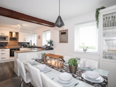 A dining area with a table set with plates bowls glasses and candles next to a kitchen with white cabinets and wooden countertops at Bywyd Da (232 Cae Du) in Abersoch