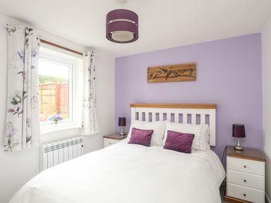 A bedroom with a double bed and purple cushions with floral curtains on the window at Bywyd Da 232 Cae Du in Abersoch