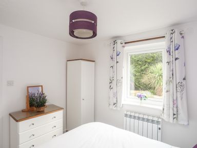 A bedroom with a white chest of drawers a tall cabinet a window with floral curtains and a purple ceiling light at Bywyd Da (232 Cae Du) in Abersoch
