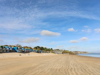 A sandy beach with a row of colorful beach huts and buildings in the background under a blue sky