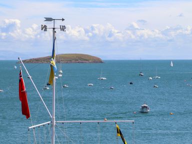 A view of a mast with flags and a weather vane overlooking a bay with several sailboats and an island in the distance at Bywyd Da 232 Cae Du in Abersoch