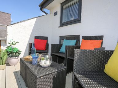 A patio with black wicker chairs with colorful cushions and a table with plants and bottles at Bywyd Da 232 Cae Du in Abersoch