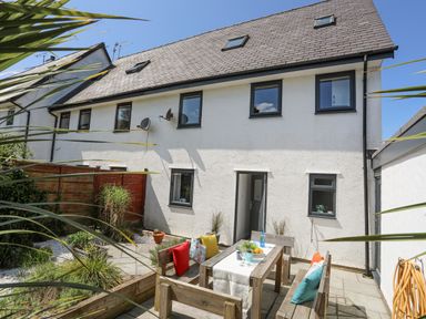 A backyard with a wooden outdoor dining table and benches with cushions beside a white house at Bywyd Da 232 Cae Du in Abersoch