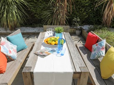 An outdoor wooden table with benches cushions plates of fruit glasses and a magazine surrounded by plants at Bywyd Da 232 Cae Du in Abersoch