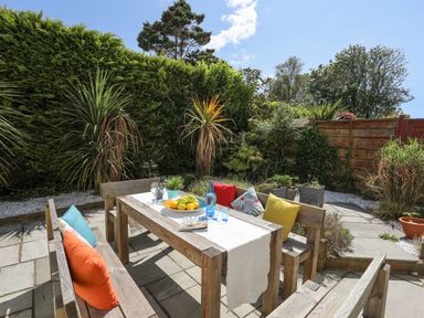 An outdoor patio with a wooden table and benches with colorful cushions surrounded by plants at Bywyd Da 232 Cae Du in Abersoch