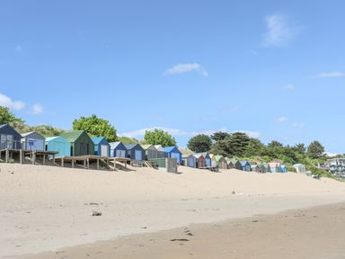 A row of colorful beach huts on raised wooden platforms along a sandy beach with trees in the background at Bywyd Da 232 Cae Du in Abersoch