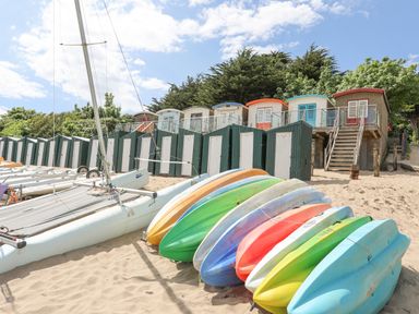 A row of colorful kayaks on sand with boats and beach huts behind at Bywyd Da (232 Cae Du) in Abersoch