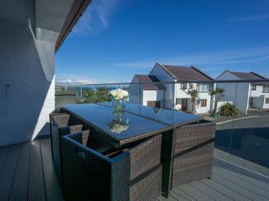 An outdoor balcony with a glass table and wicker chairs overlooking houses at Cromlin Glas in Abersoch