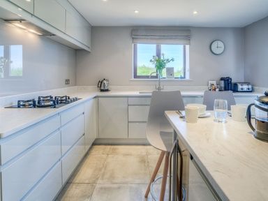 A kitchen with white cabinets a gas stove a window with flowers and a dining table with chairs at Cromlin Glas in Abersoch