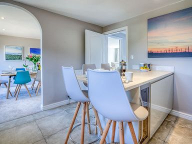 A kitchen island with white bar stools and cups next to a dining area with a table, chairs, and flowers at Cromlin Glas in Abersoch