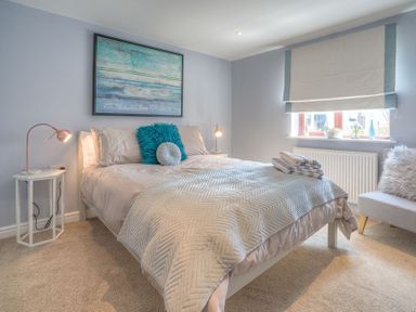 A bedroom with a bed covered with pillows and a blanket bedside tables with lamps a window with blinds and a chair at Cromlin Glas in Abersoch